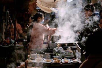 一位戴口罩的妇女在街头小吃摊上烹饪食物 热气弥漫 顾客在旁