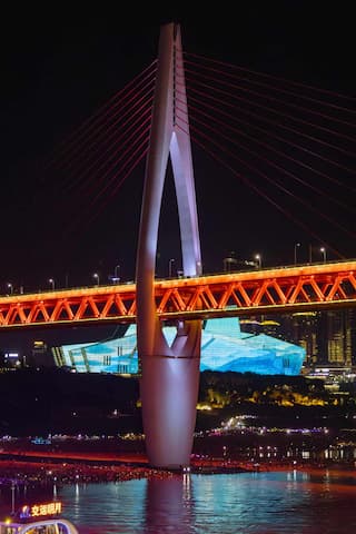 夜间拍摄的桥梁主体结构红色桥身和连接桥塔的缆绳城市建筑在背景中水面有倒影