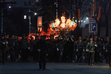 一群人夜间抬着灯笼在街道上行进队伍穿着相似的服装街道两旁有树木和建筑物