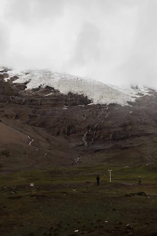 图片展示了被云雾笼罩的雪山，山坡上有水流形成小瀑布，山脚下是绿色的草地，有零星的人影。