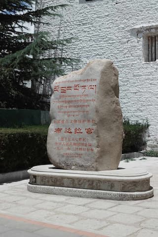 图片中，一块刻有藏文和汉字的石碑立于布达拉宫附近，石碑放置在雕刻的基座上。