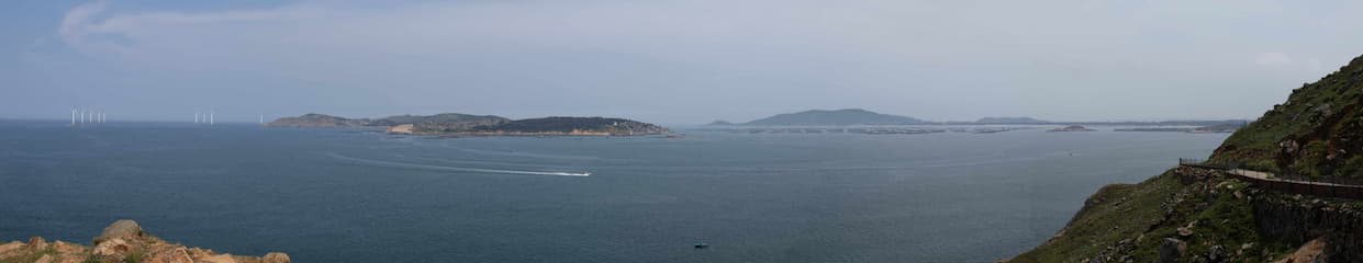 海面平静远处有岛屿和风力发电机近处是山坡和道路