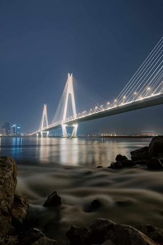 夜幕下，一座桥梁横跨水面，桥身灯光照亮夜空，水面波光粼粼，岸边有岩石
