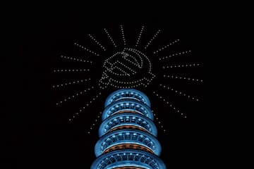 夜空中，无人机灯光组成了镰刀锤子标志，下方是一座多层塔建筑。