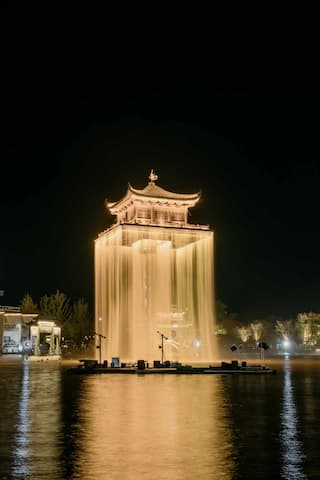 夜间拍摄的塔楼被灯光照亮，四周环绕着水幕，水面平静地倒映着塔楼的景象。