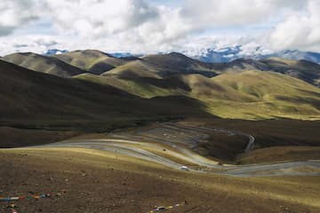 图片展示了高原地区的山地景观，蜿蜒的道路穿梭于山峦之间，远处的雪山在云层下若隐若现。