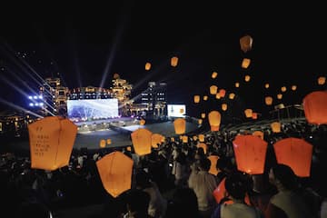 夜空中漂浮着许多孔明灯，下方是人群和灯光照亮的建筑物。