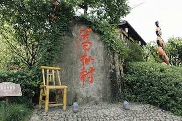 图片展示了一个乡村的角落，一块刻有文字的石头立于绿植之中，旁边放着一把椅子。