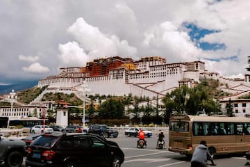 图片展示了布达拉宫的远景，前景有车辆行驶，天空中有云。