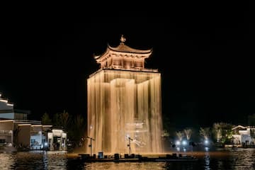 图片展示了一座被灯光照亮的塔楼，塔楼周围环绕着喷泉，水流如瀑布般倾泻而下。塔楼和喷泉倒映在平静的水面上。
