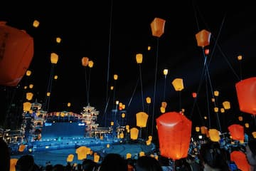 夜空中，许多橙色的天灯在漂浮，下方有建筑物和人群。